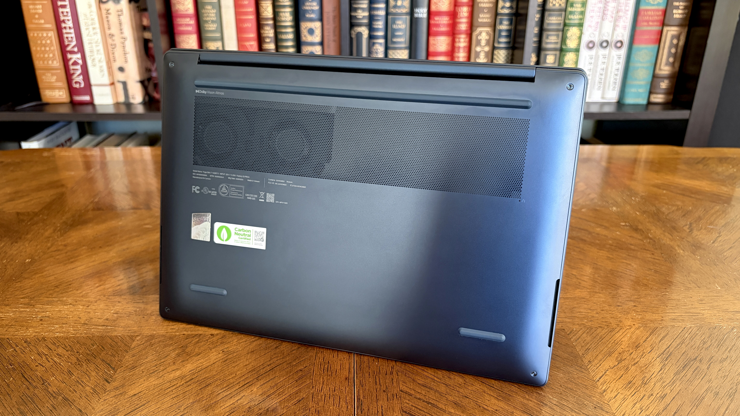 A laptop viewed from the bottom, showcasing cooling vents and certification stickers, is placed on a wooden table, with a bookshelf full of books in the background.