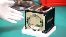 closeup photo of a small cubical brown satellite sitting on a green table, with a white-gloved hand next to it for scale