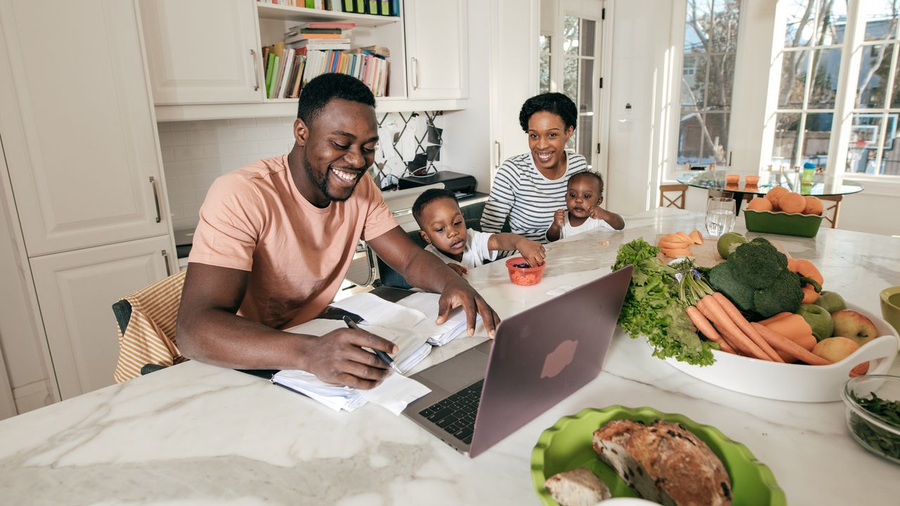 Dad works on financial paperwork at the kitchen island while Mom and their two kids watch.