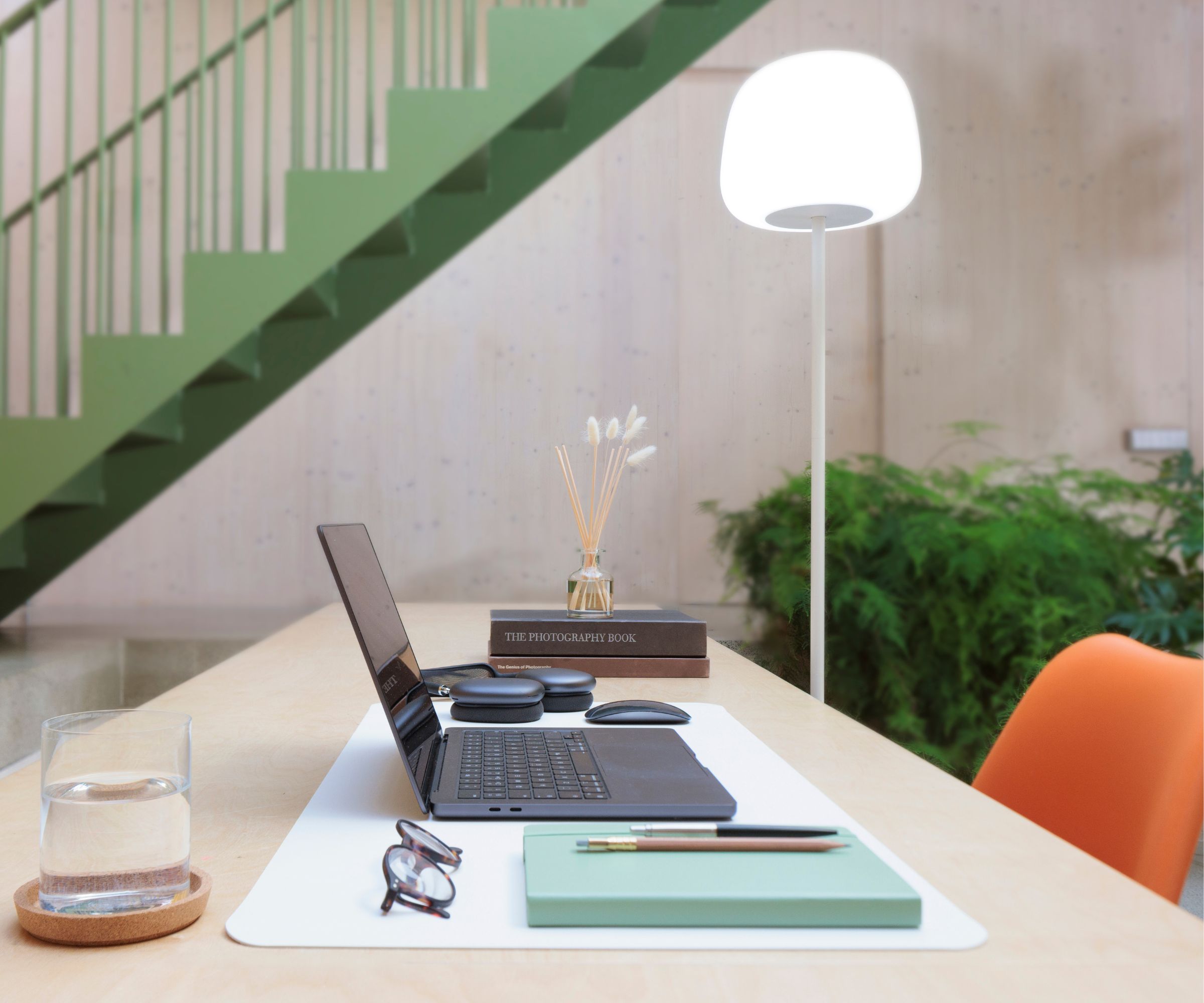 Desk with laptop, notebook, a cup of water, and an orange chair tucked underneath, with the Govee floor lamp beside it and a green staircase and plants in the background.