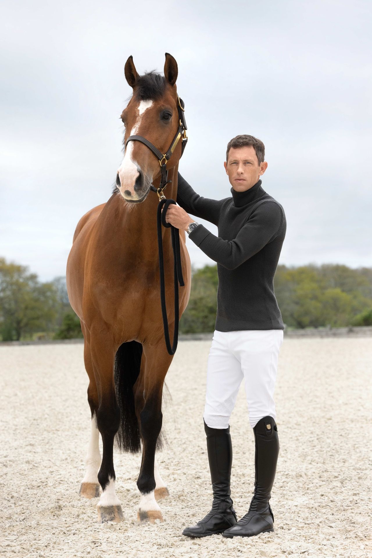 Scott Brash stands next to a tall chestnut horse in white jodhpurs and a black roll neck