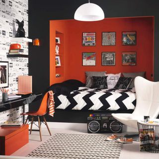 Retro black and orange bedroom, painted black and orange walls, bed built into alcove raised on platform, framed wall mounted album covers, white wing backed chair large ghetto blaster, black gloss desk, wallpaper with cassette tape print, white glass pendant light