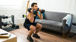 Man performs squat in his living room, holding dumbbells by his shoulders