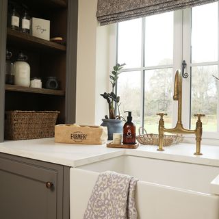 Country-style kitchen sink in front of a window, with brass taps, white worktop, and grey cupboards