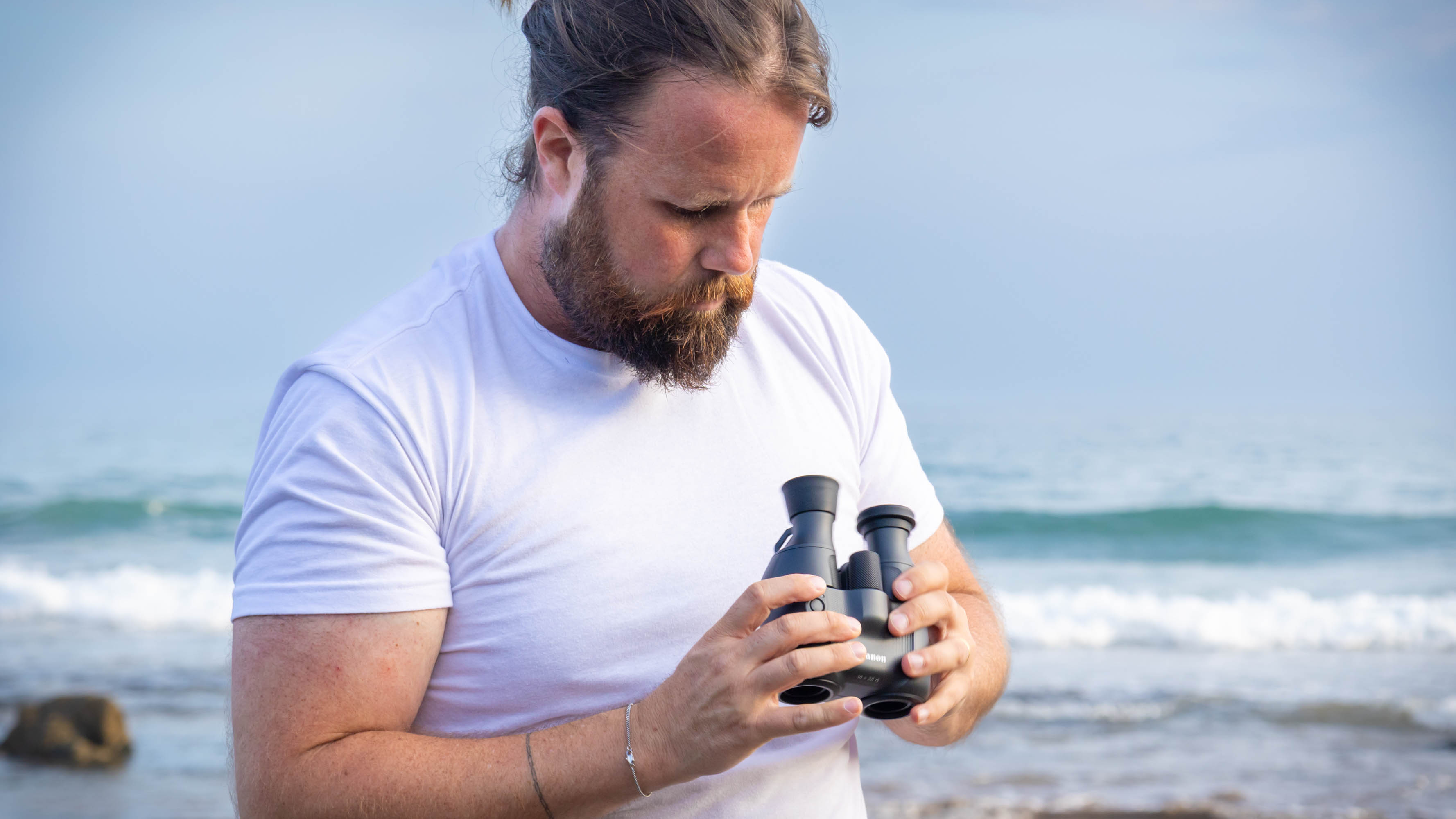 A male looking at the Canon 10x20 IS binoculars on a beach, with the sea behind them.