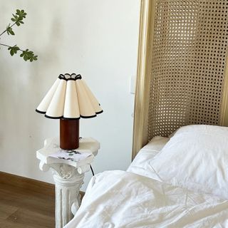 A black and white pleated lampshade on a wooden base. The lamp is on a column by the white and gold bed