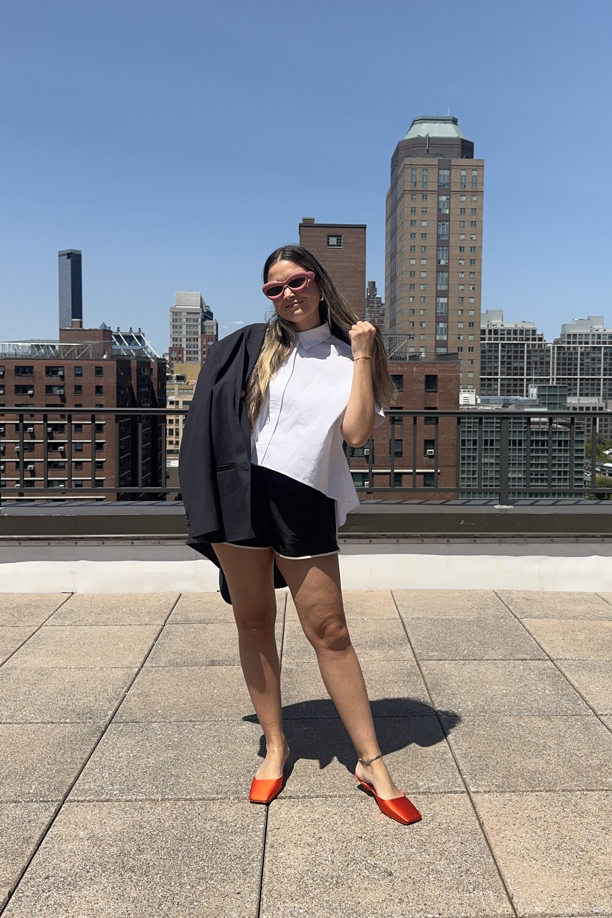 Writer Irina Grechko in black track shorts with a white short-sleeve button-down, black blazer, orange mules, and pink sunglasses.