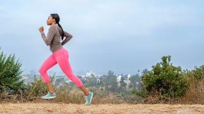 woman in pink leggings and grey sleeved top running sideways to the camera on a dusty track and shrubs beside her and blue sky behind