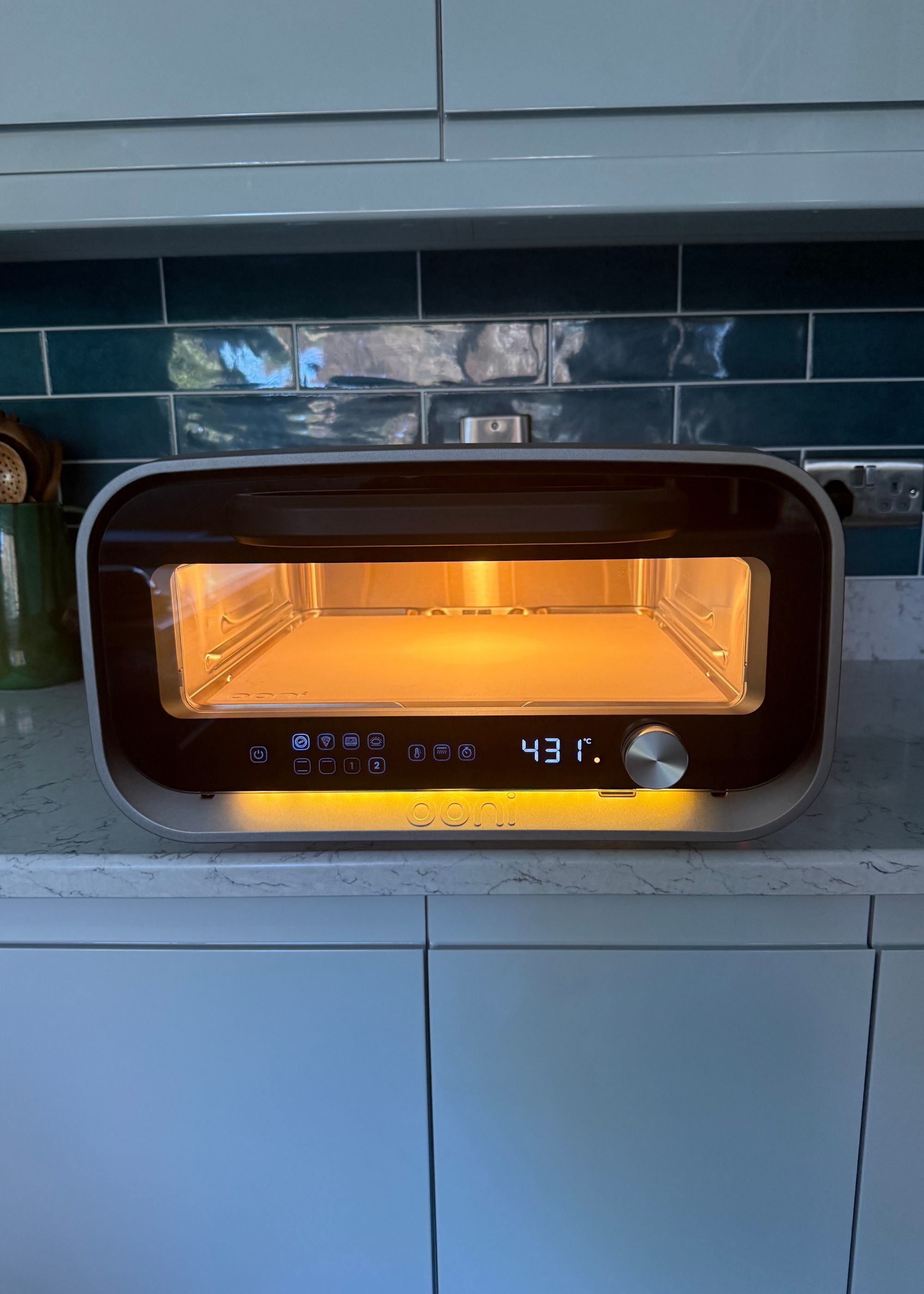 Ooni's volt 2 indoor pizza oven with the oven light on and a reading of 431 degrees on the panel, sitting on a countertop with a tiled teal backsplash behind and light gray cabinets above and below it