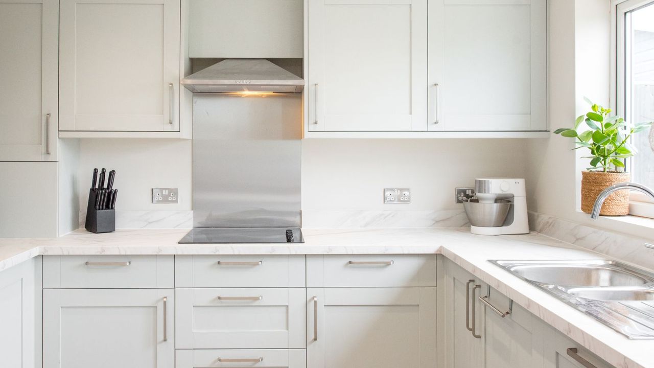 A general interior view of a newly fitted sage green, grey shaker style kitchen with stainless steel handles, extractor fan hood, splash back, oven, knife block, food mixer, potted plant, recessed ceiling spot lights and marble effect worktop within a home