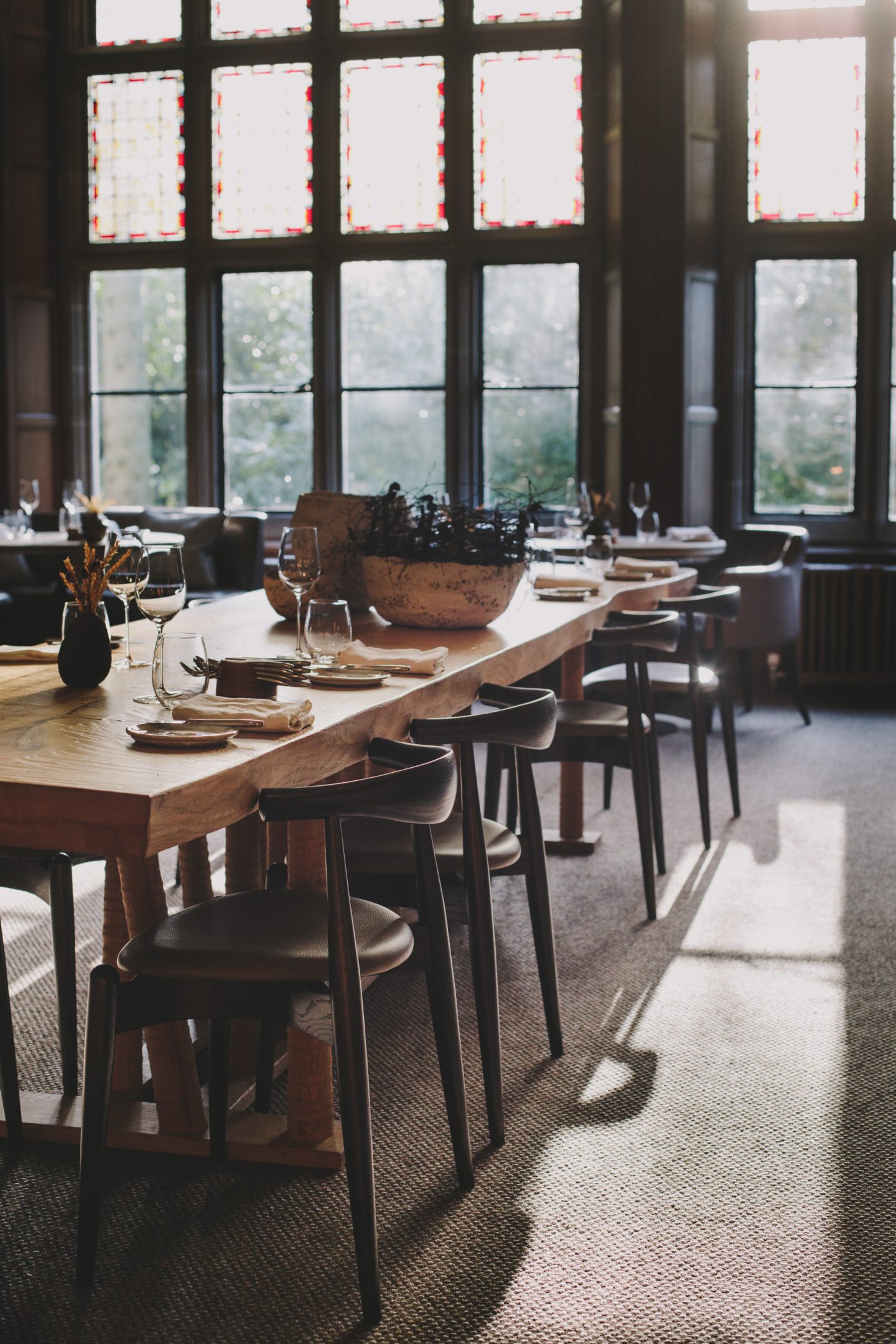 A modern rustic hotel restaurant features a large wooden table with dark wood and leather chairs, crafty accessories, and dried flower bunches.