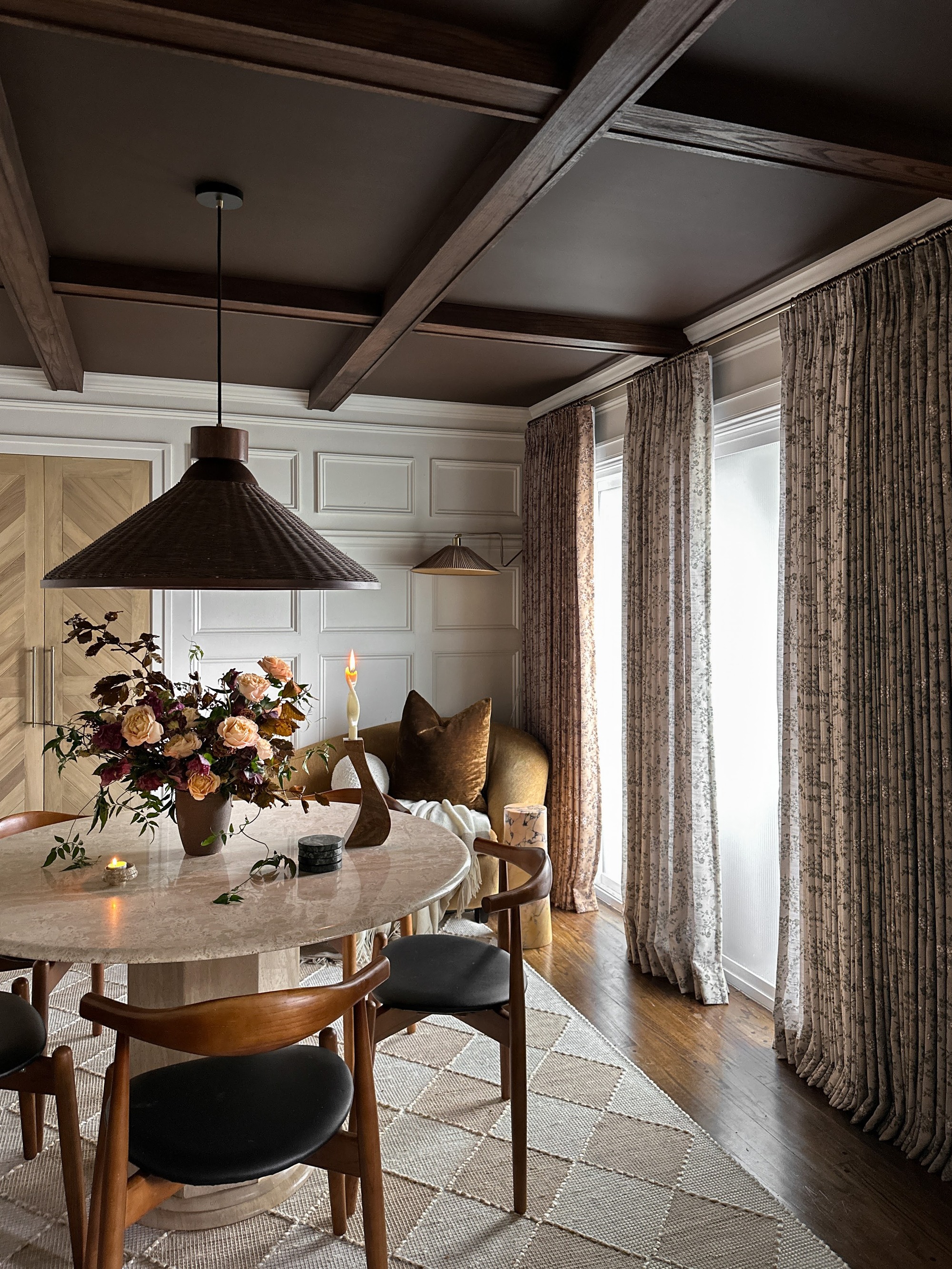 A modern dining room with a dark color palette and traditional touches. The walls are white, the ceilings are a dark brown, and the floors are hardwood with a jute rug. There is a round, beige marble dining table with wooden and black leather dining chairs. There are windows with glazed film and curtains on them.