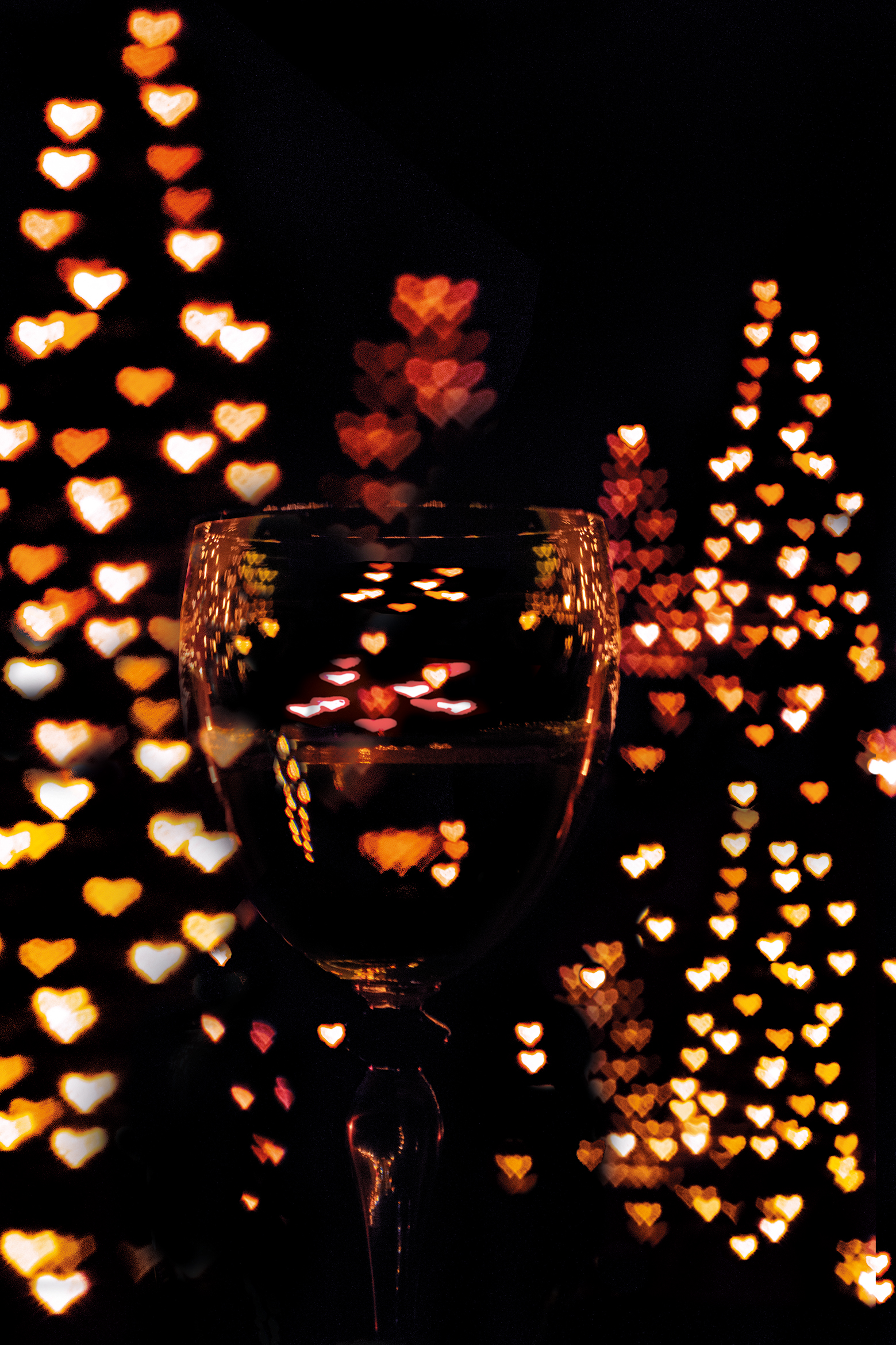 A wine glass in focus, reflecting warm heart-shaped bokeh lights from colorful Christmas trees in the background against a dark backdrop