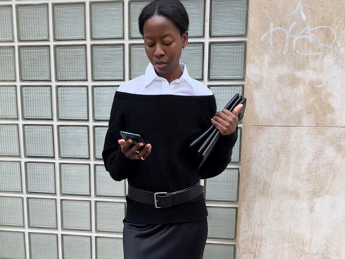 Paris-based fashion influencer Sylvie Mus stands on the sidewalk while looking at her phone wearing a black off-the-shoulder top layered over a white button-down shirt along with clutch bag, belt, and satin skirt