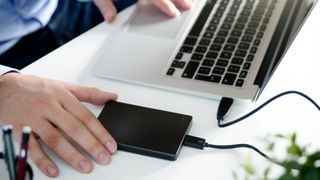 A man's hands and partial body pictured at a desk. He's using a Mac with an external hard drive plugged in.