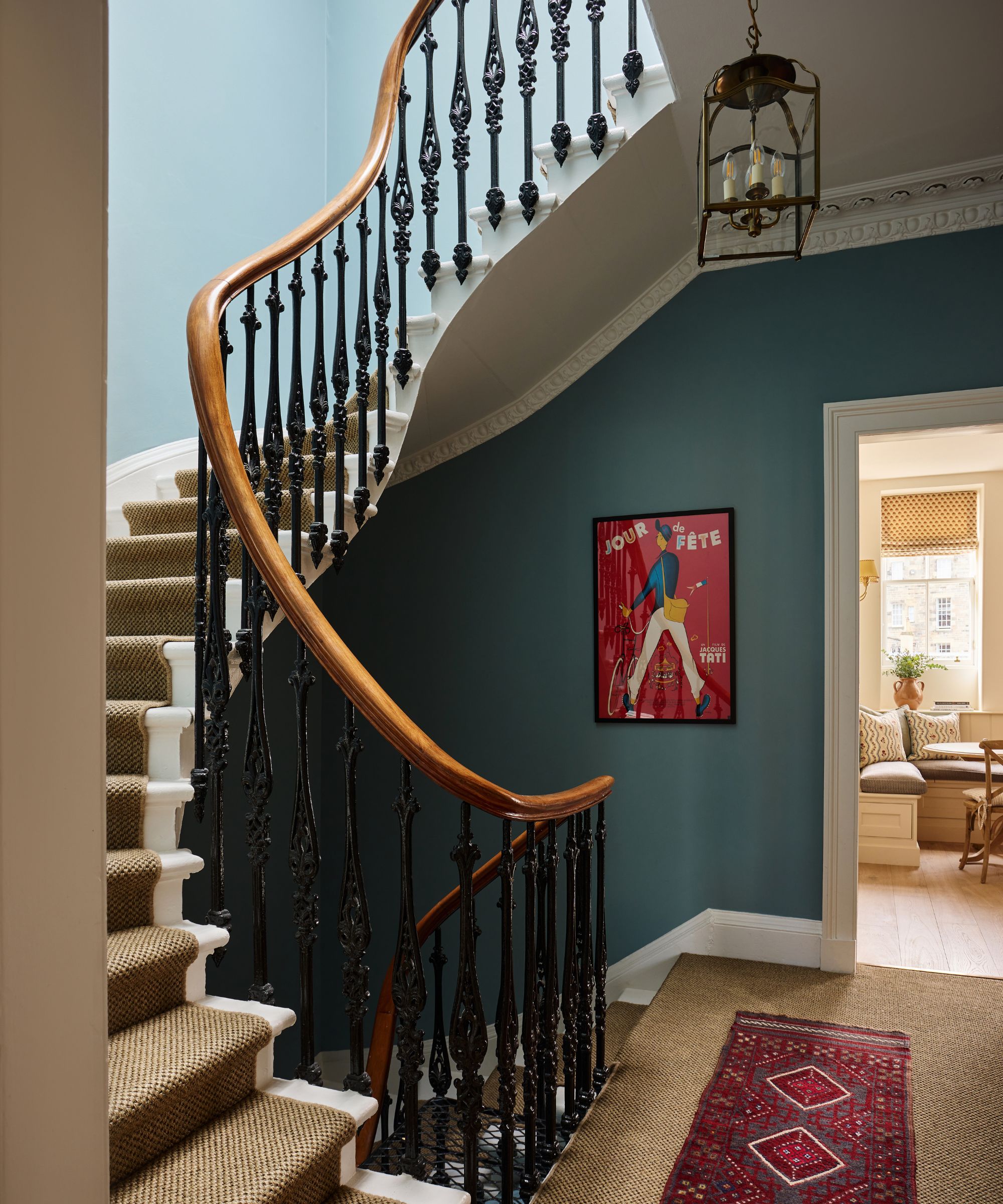 Elegant entryway with a sweeping curved staircase, black wrought-iron balusters, deep teal walls, and a colorful framed vintage poster