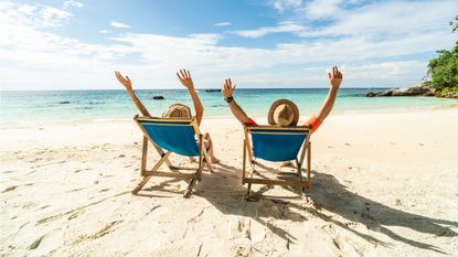 An older couple celebrate with their hands in the air while sitting in beach chairs on the beach facing the ocean.