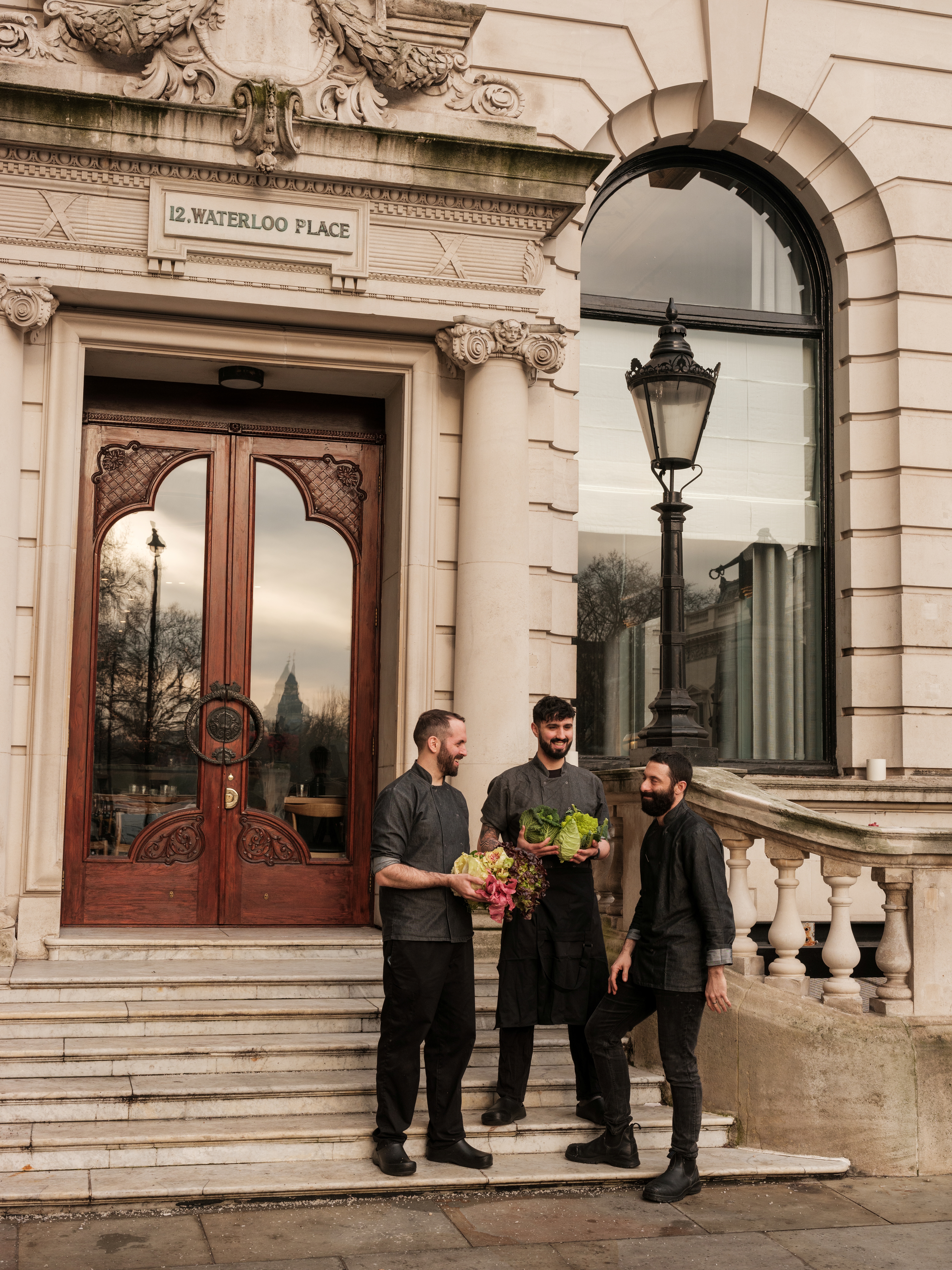 Three young chefs dressed in dark gray and black cooking attire stand joyfully in front of the entrance to a stone-clad, historical building with a tag reading "12 Waterloo Palace", ornate wooden doors, and a vintage light pole.
