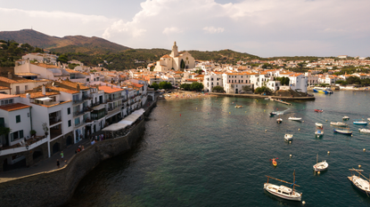 Coastline of Costa Bravo. White buildings with orange roofs meet the blue water with boats sailing across and a green hill landscape in the background