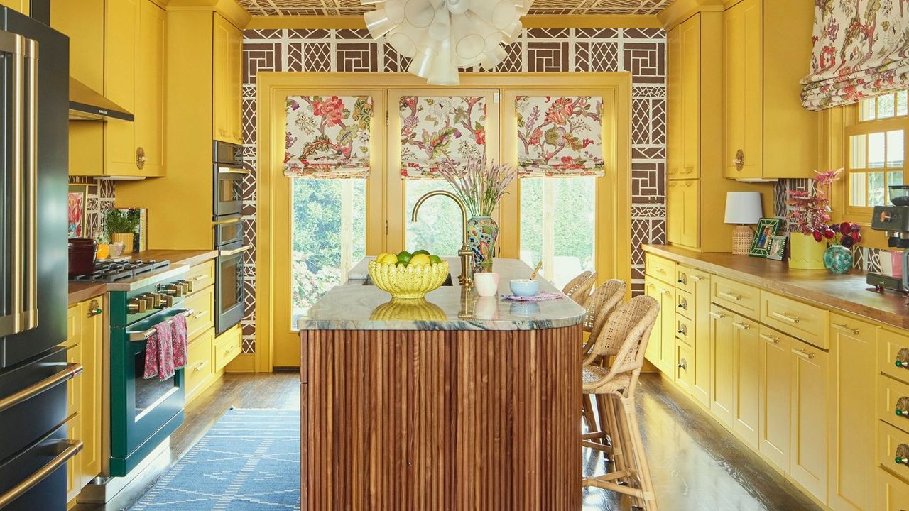 Bright yellow kitchen with wallpaper on ceiling, floral blinds, marble island and a dark green fridge and oven 