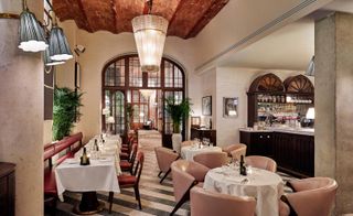 A restaurant with decorated tables, chairs, a bar, potted plants, a large chandelier, wall lights and large doors.