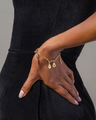 The hand of a model wearing pink nail polish and a golden bracelet with avocado-shaped charms is captured in a photograph.