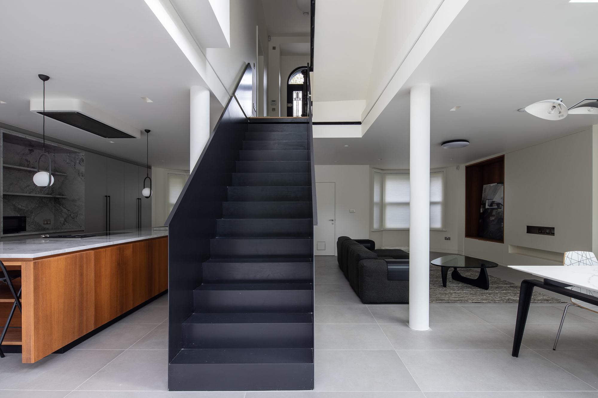 A minimalist open plan kitchen with a wooden island, stone tiles, and a black staircase in the center