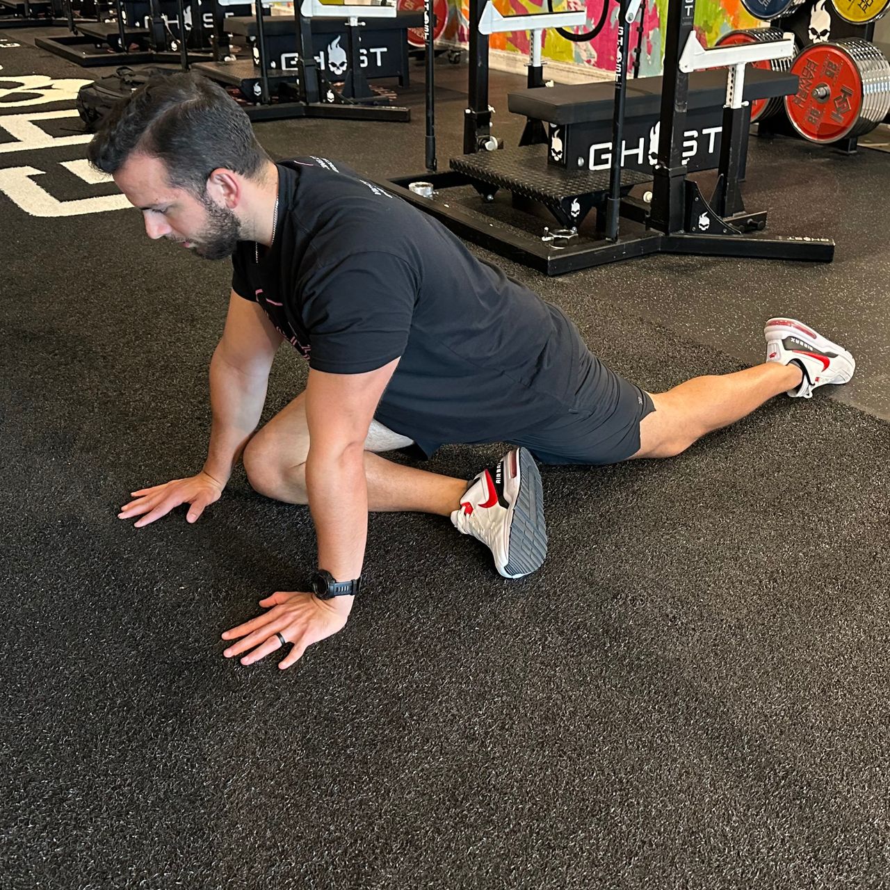 An employee at Vice City Sports Medicine performs a pigeon pose in a gym setting. They are on the floor, with their right shin horizontal to their body, hands on the floor, arms straight, and left leg behind them.