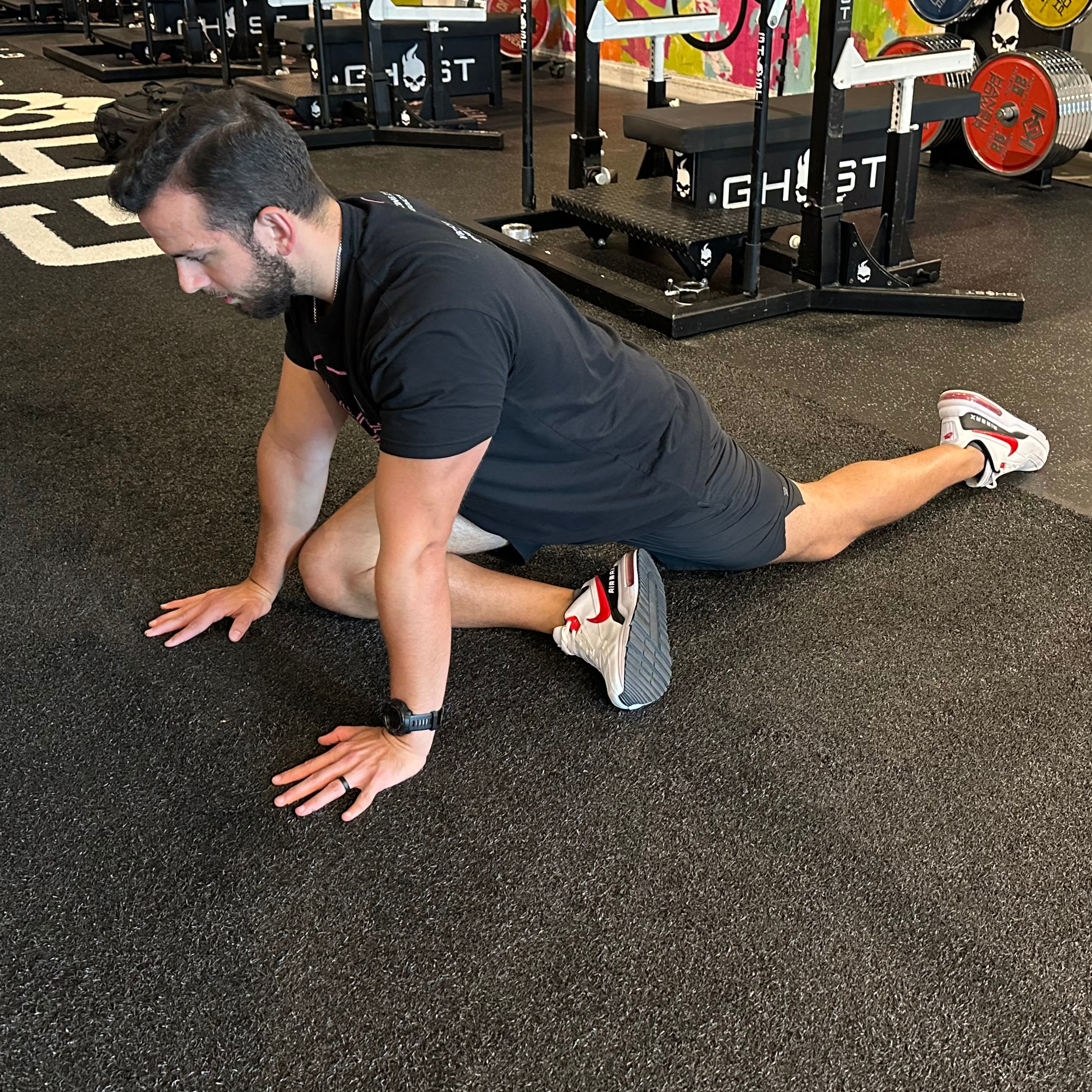 An employee at Vice City Sports Medicine performs a pigeon pose in a gym setting. They are on the floor, with their right shin horizontal to their body, hands on the floor, arms straight, and left leg behind them. 