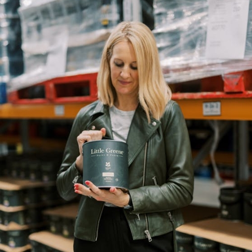 Image of a woman in a gray top and green leather jacket holding a can of paint and looking at it.