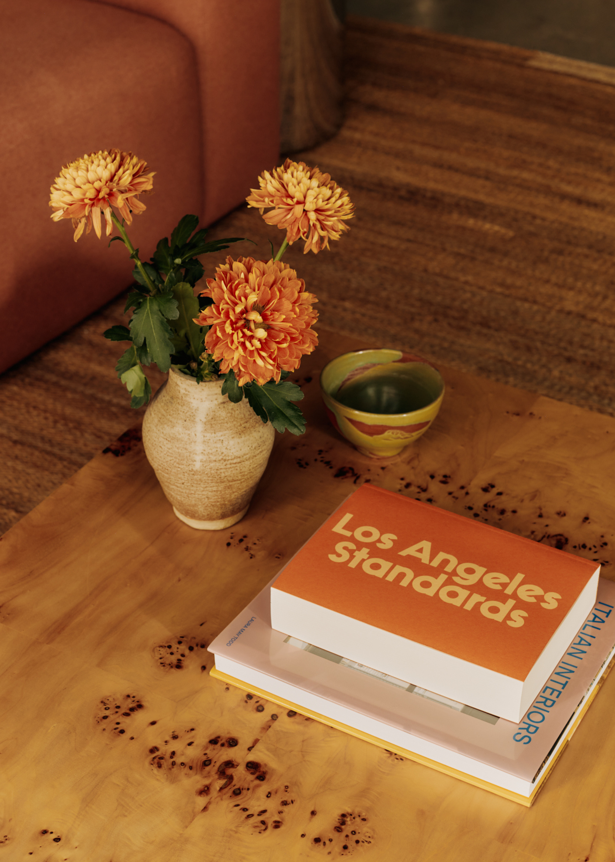 A burl wood coffee table styled with a small vase of fresh orange hued flowers and two books including an orange LA Standards book next to a small green bowl