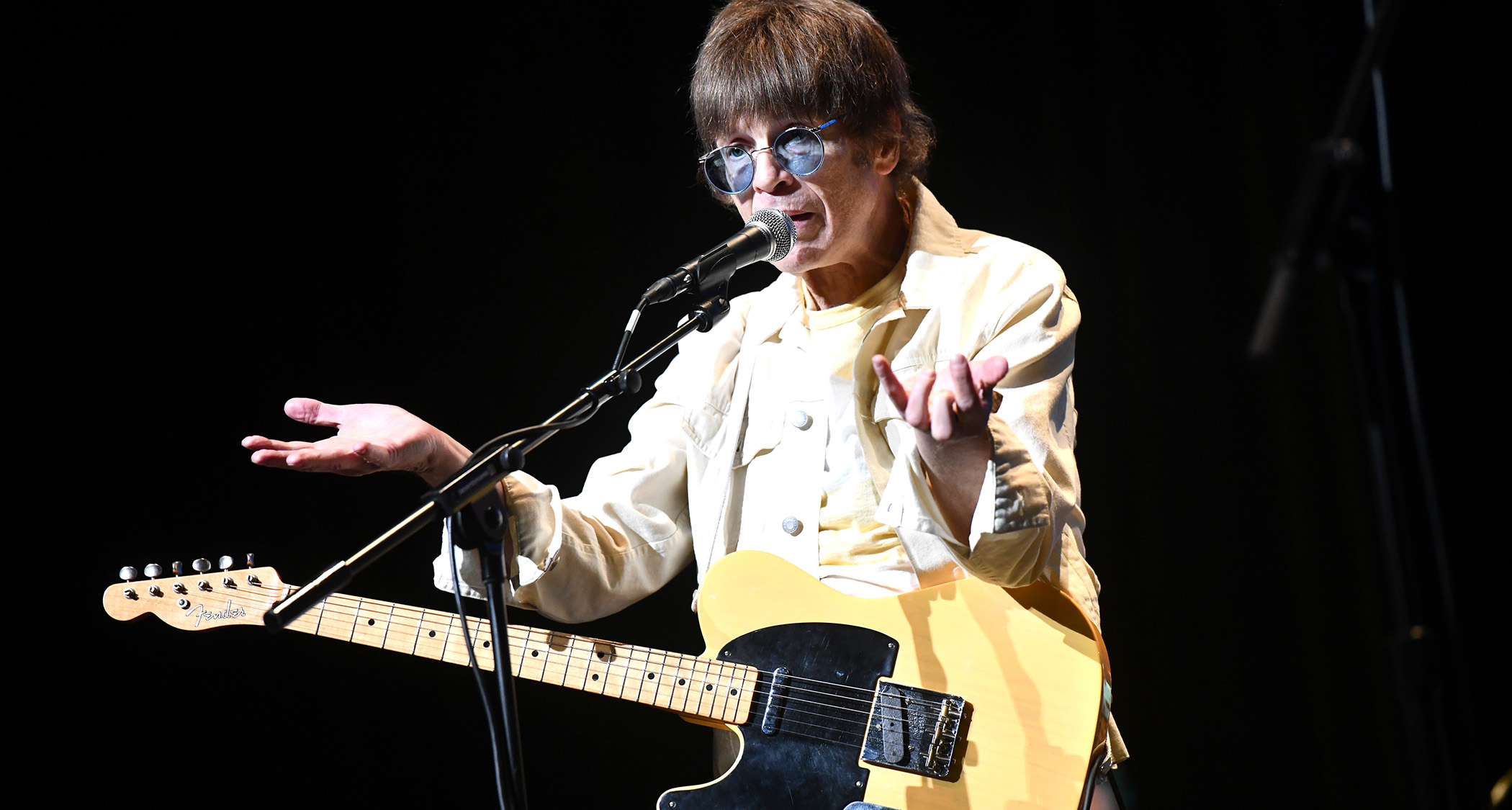 Elliot Easton wears round sunglasses with a pale blue tiny and holds his hands out as he addresses the crowd. He has a Butterscotch Blonde Telecaster.