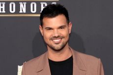 A headshot of Taylor Lautner wearing a brown suit jacket looking at the camera and smiling