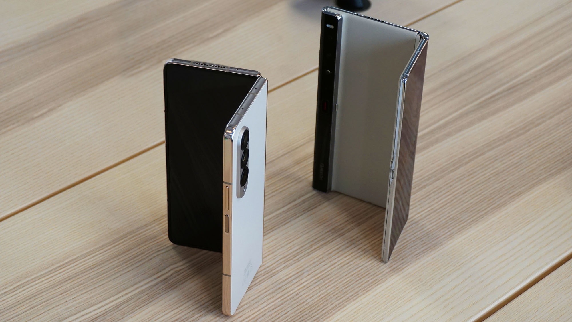 A partially folded Samsung Galaxy Z Fold 4 next to a partially folded Huawei Mate Xs 2, both on a pine table