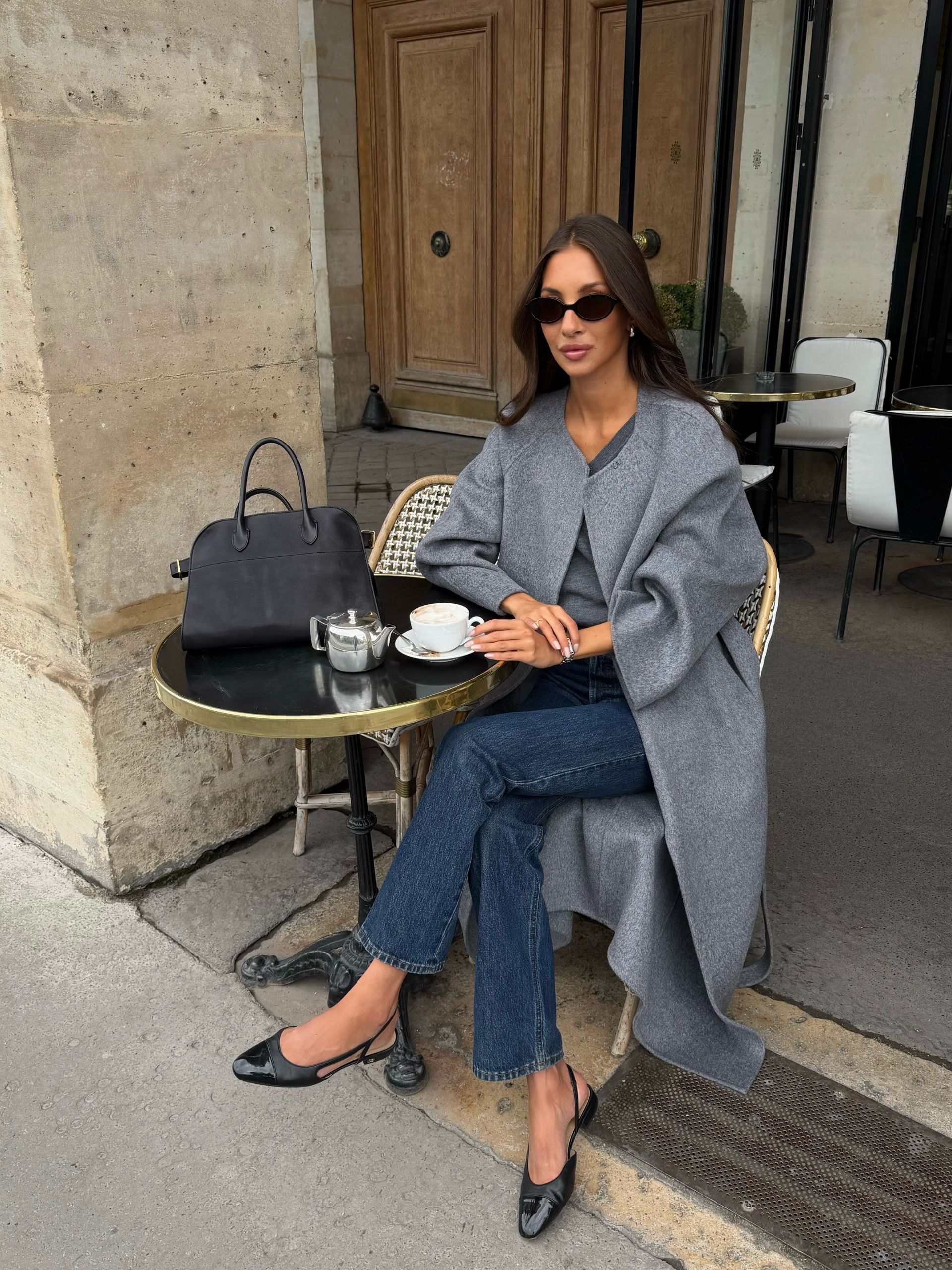 An image of an influencer in Paris wearing a collarless grey coat, dark-wash jeans and ballet flats sitting at a cafe