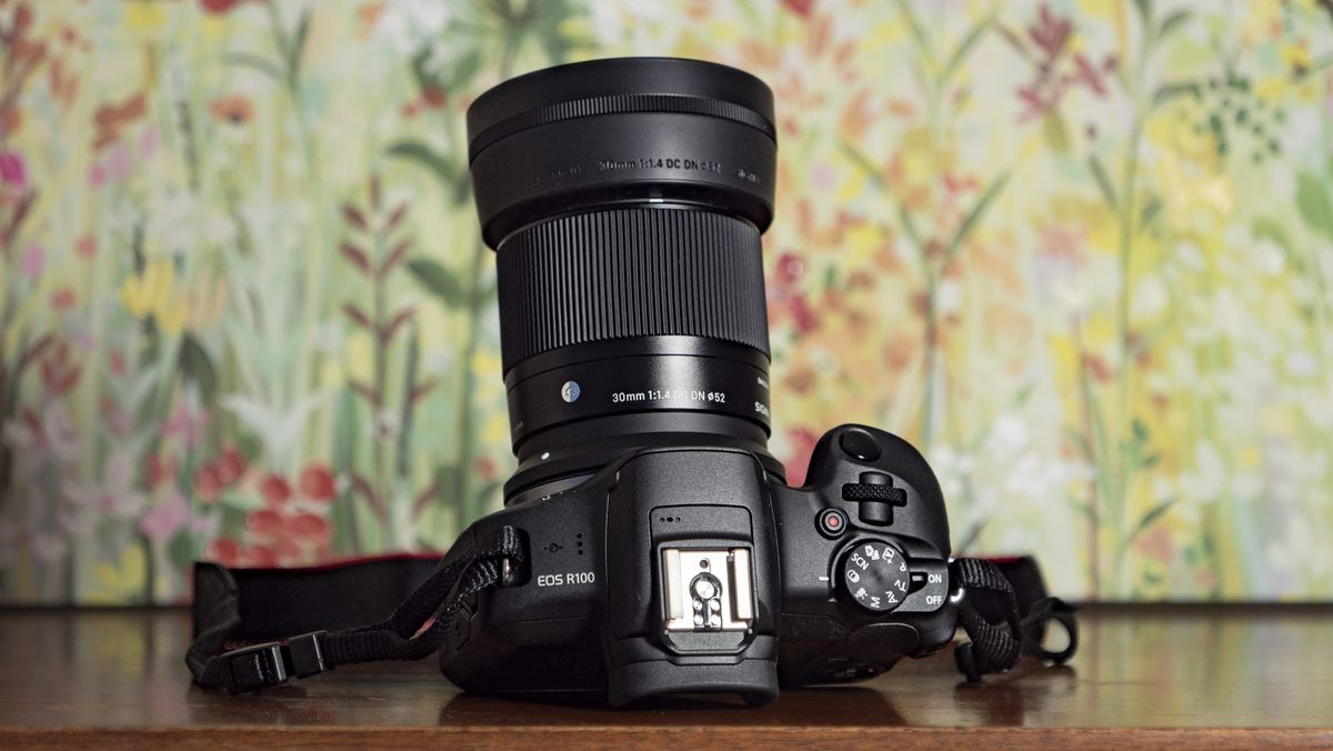 Sigma 30mm F1.4 DC DN Contemporary lens attached to a Canon EOS R100, on a wooden surface with floral backrop