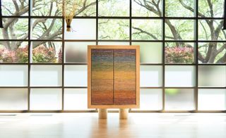 Daytime image, wall of small black framed windows, bottom rows opaque glass, tapestry in a light wooden frame stood on a glossed wooden floor, outside view of trees, flowers and shrubs