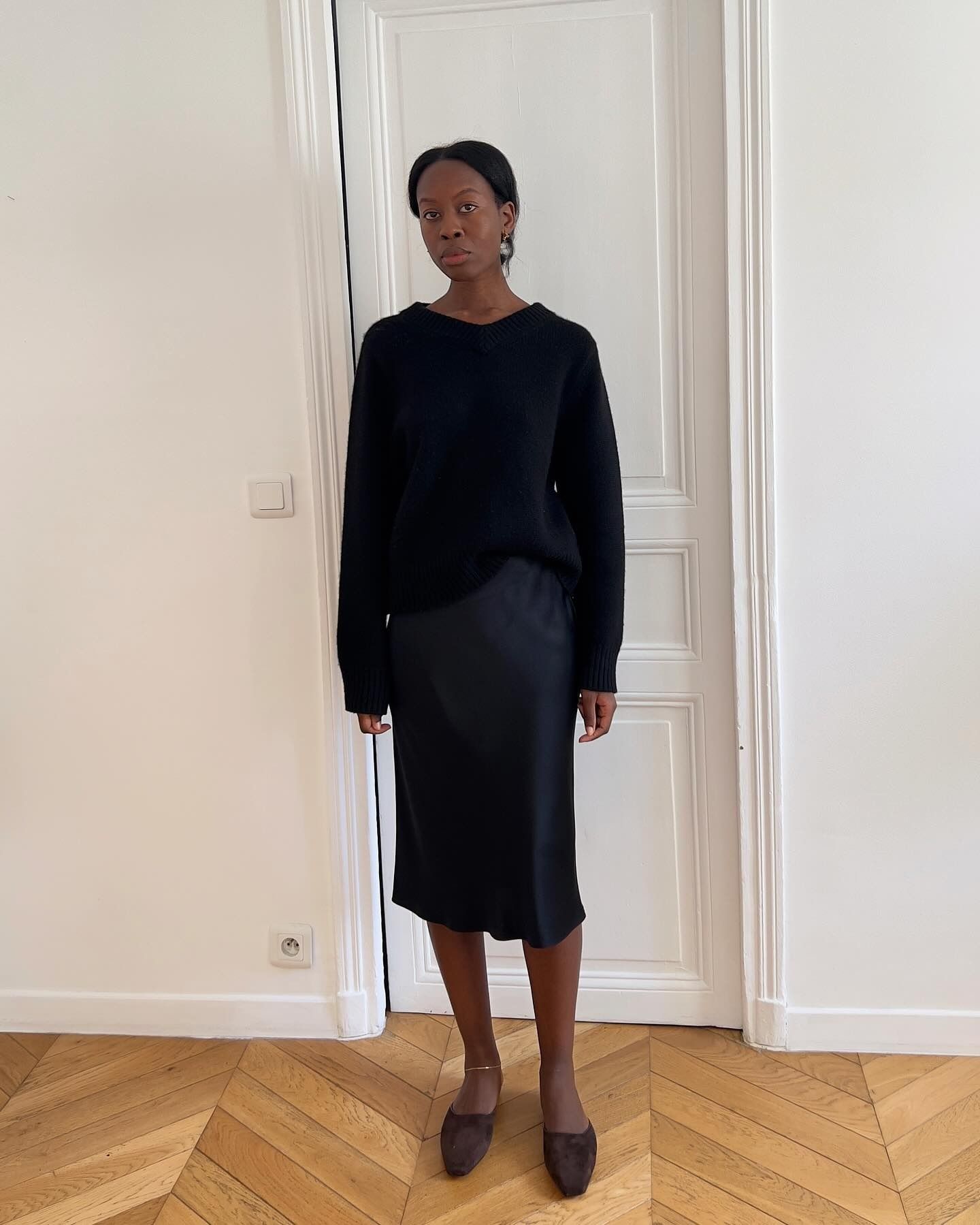 Sylvie Mus wearing a V-neck black sweater, navy-blue silk slip skirt, and brown suede mules.