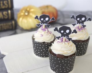 Halloween themed chocolate cupcakes with black and white spotted cases, white vanilla frosting, purple sprinkles, skull cupcake toppers with wooden sticks and spray painted pumpkins in background