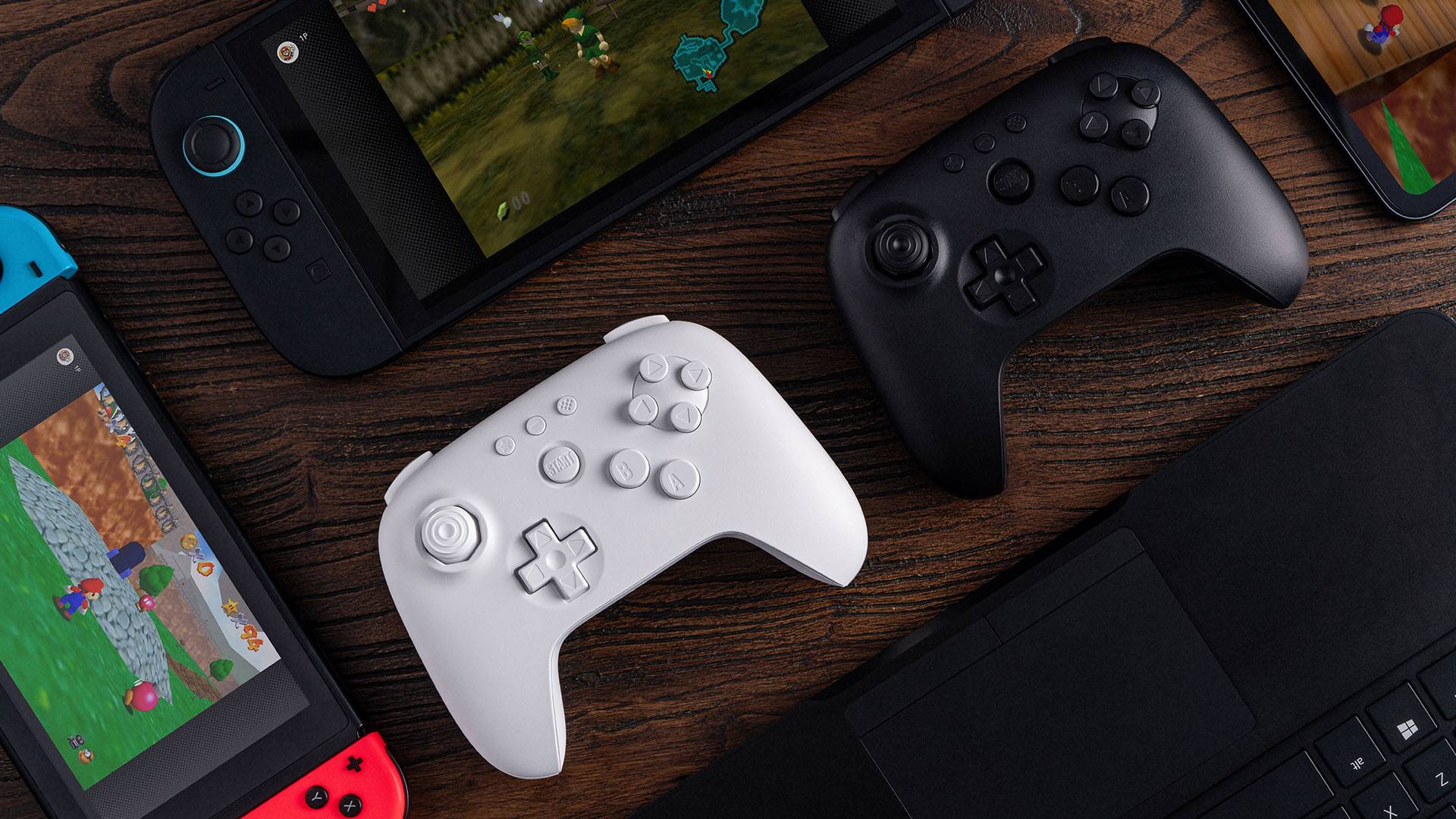 8bitdo N64 controllers in white and black surrounded by Switch 2, original Switch, and gaming laptop on woodgrain desk.