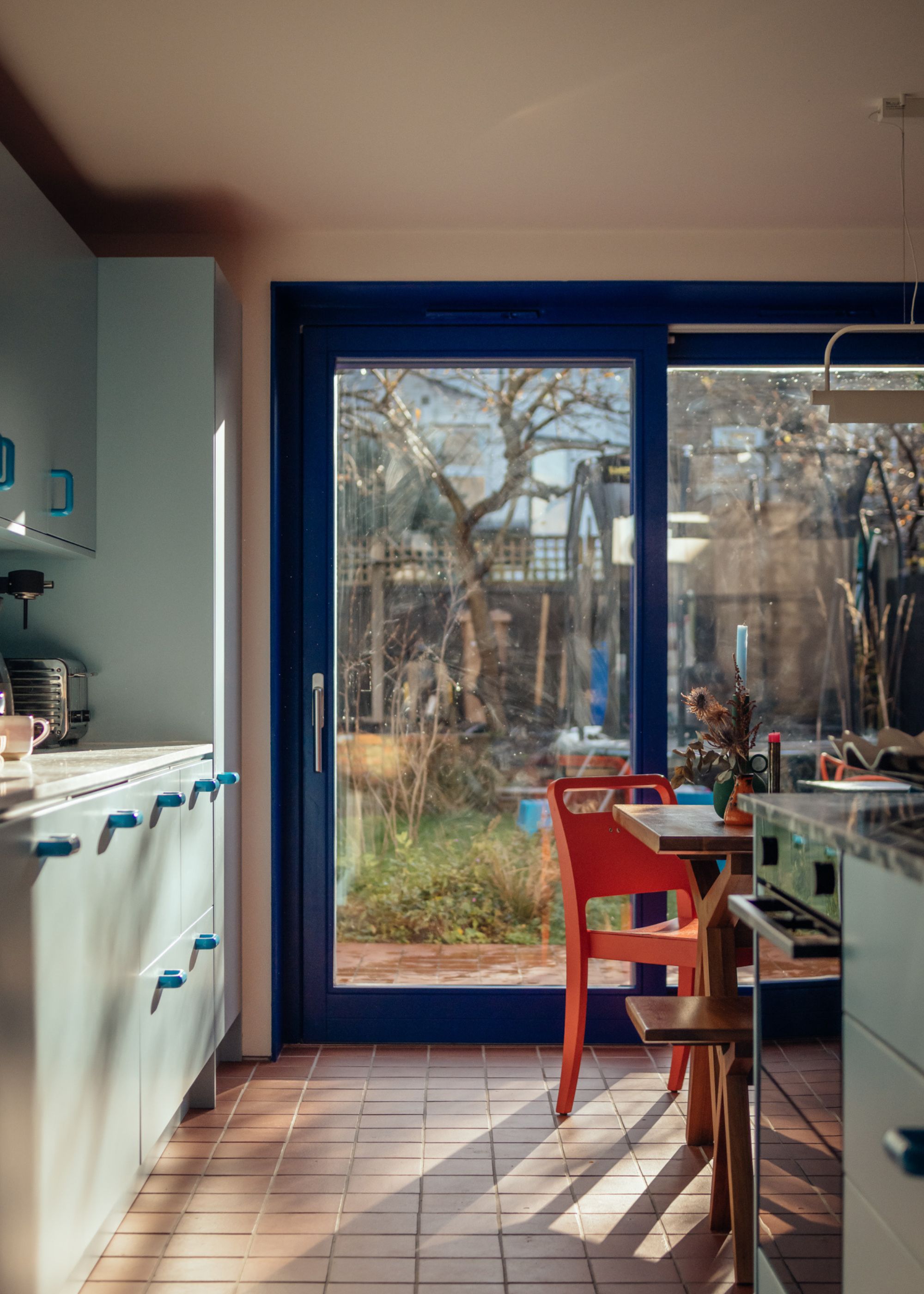 A modern blue kitchen extension with pale blue cabinetry and deep blue sliding doors leading to the garden with a vibrant orange dining chair by the wood table