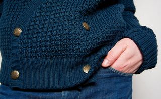 Close up a man wearing a knitted blue cardigan with brass buttons, top of jeans with his hand half way in the pocket, white backdrop