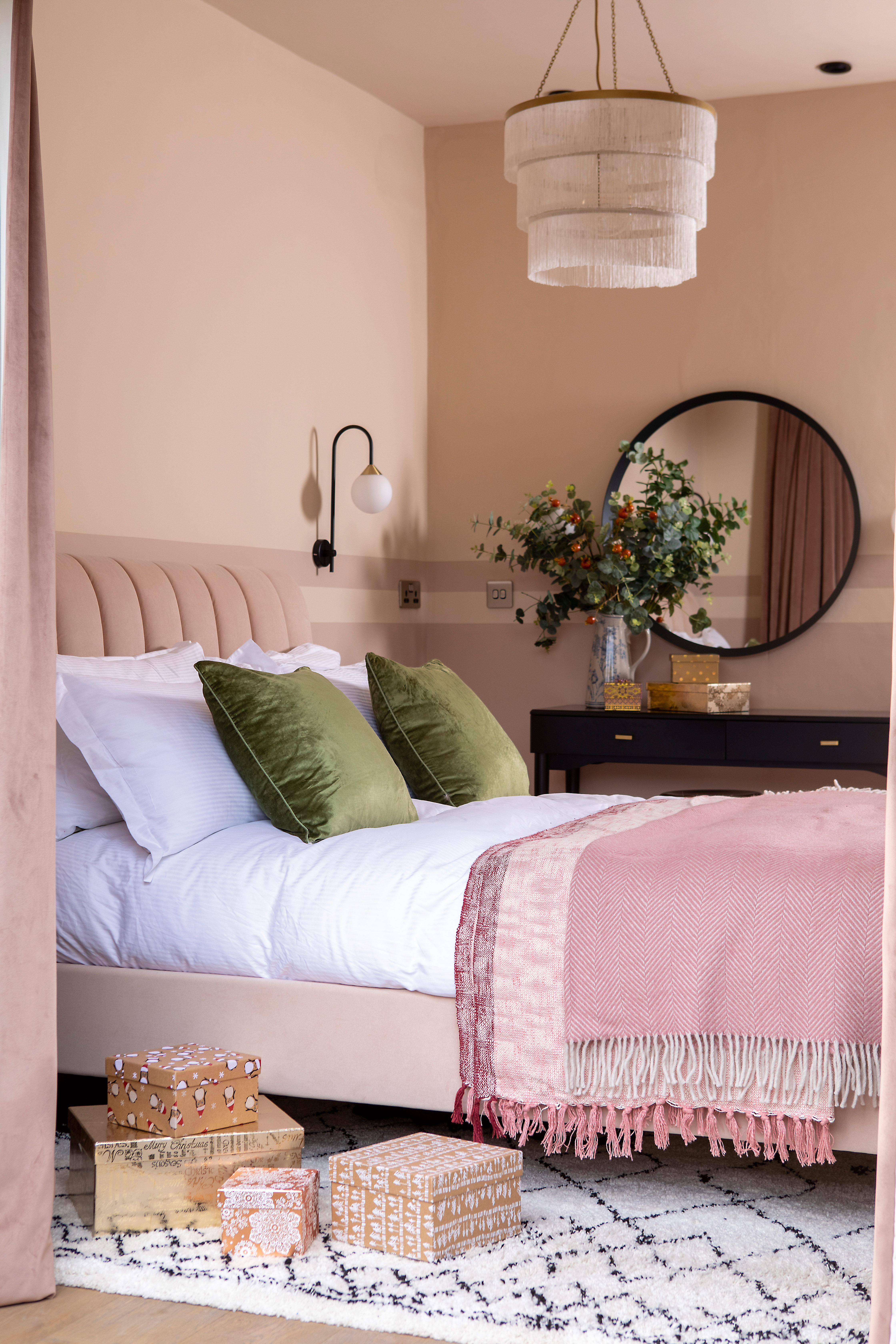 A bedroom with peach wall paint decor, with green cushions, black framed round mirror and statement pendant ceiling light with fringed lampshade