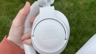 Någon som håller upp JBL Tune 750BTNC-hörlurarna i vitt nära kameran med JBL-logotypen på ena öronkåpan.