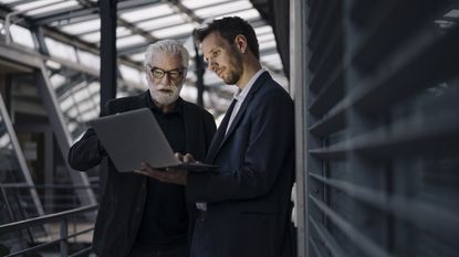 An older man and a younger man discuss selling the family business while looking at a laptop.