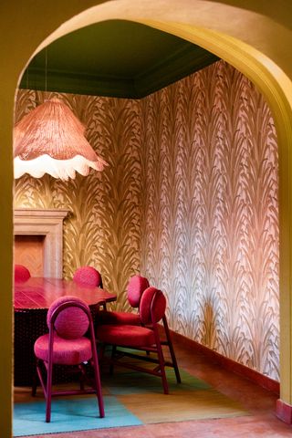 A colorful dining room with tropical leaf patterned wallpaper in a beige and light pink colorway. There is a pink-stained wooden dining table with modern magenta dining chairs around it. The rug below is turquoise and there is an oversized pendant light overhead.