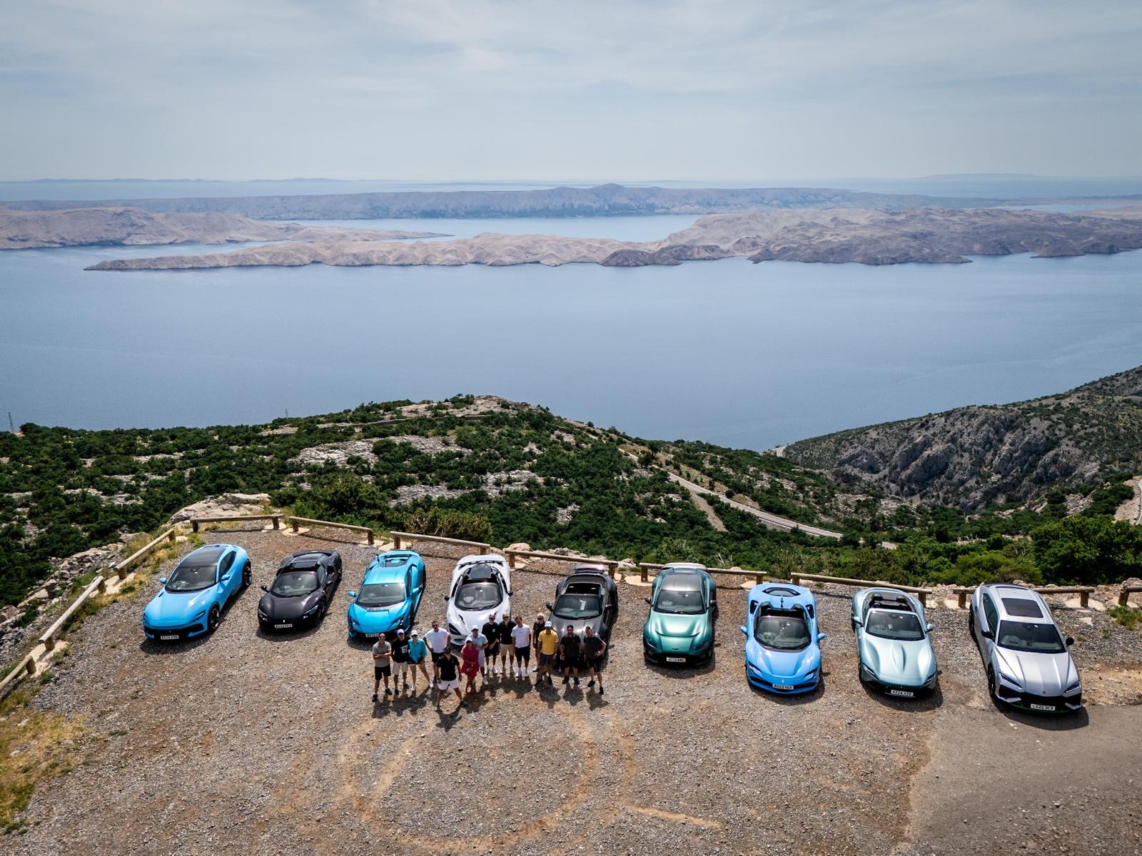 A group of supercars parked on the edge of a cliff in the Croatian coast. The drivers are standing in front of the cars.