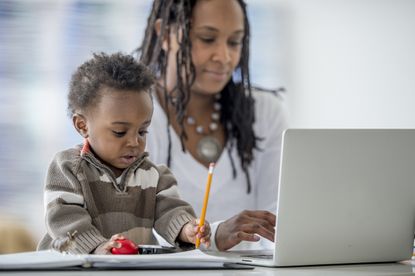 A woman of African descent is at home with her baby boy. She is working from home on her laptop. Her son is playing with a pencil and toy car beside her.