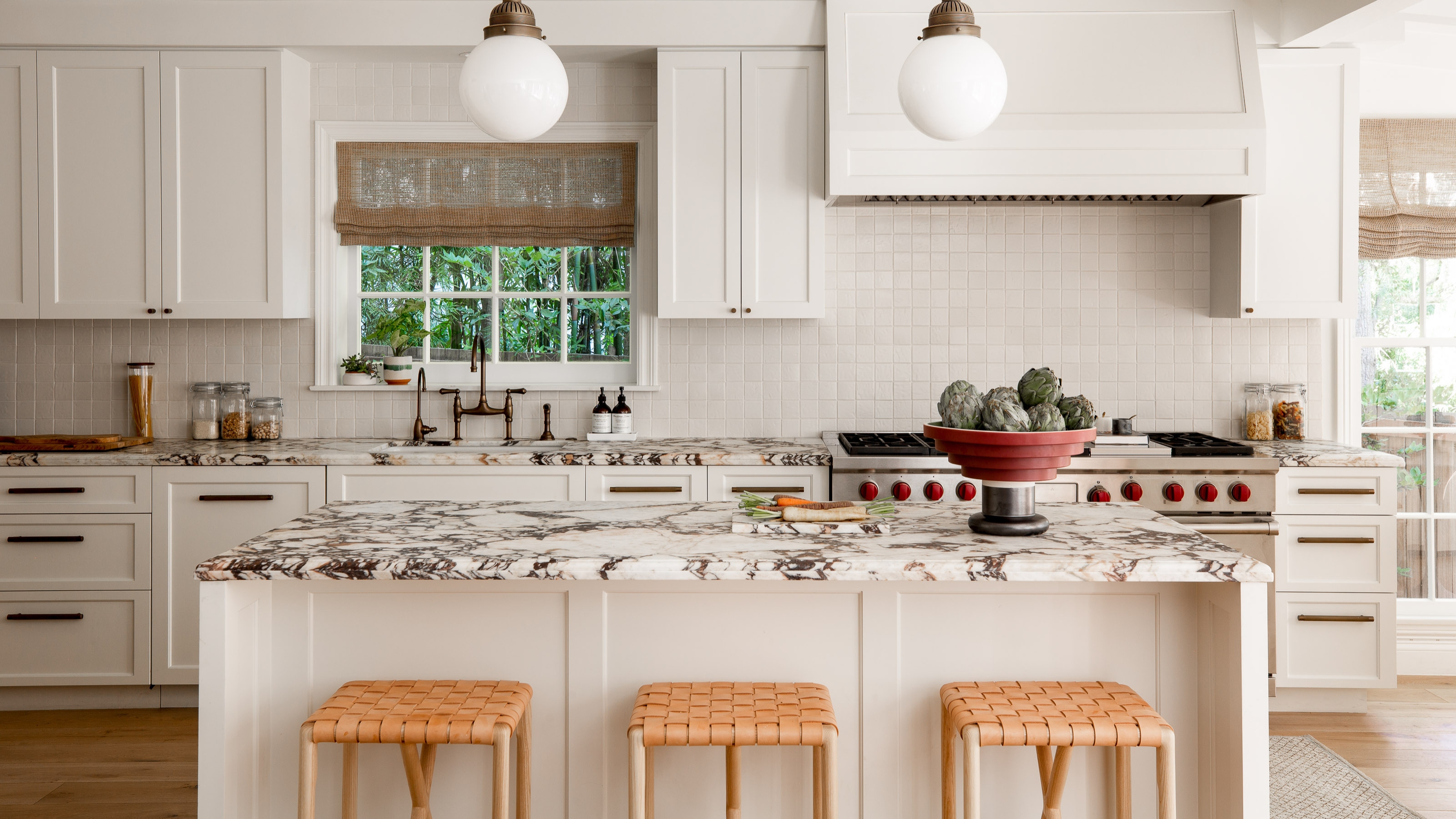 Image of a spacious white kitchen with white cabinetry and a white and dark gray marble countertop. There is a large island in the middle with three barstools that have a leather basket weave top.