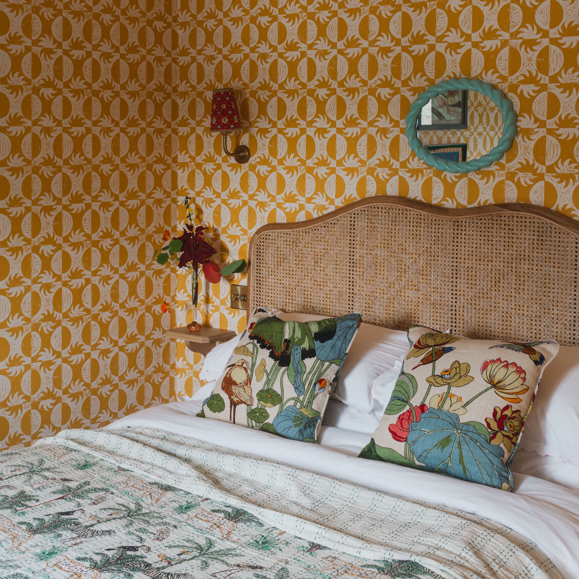 bedroom with patterned yellow wallpaper, double bed with wicker headboard and colourful cushions, blue frame mirror on wall and wall lights with pleated lampshades